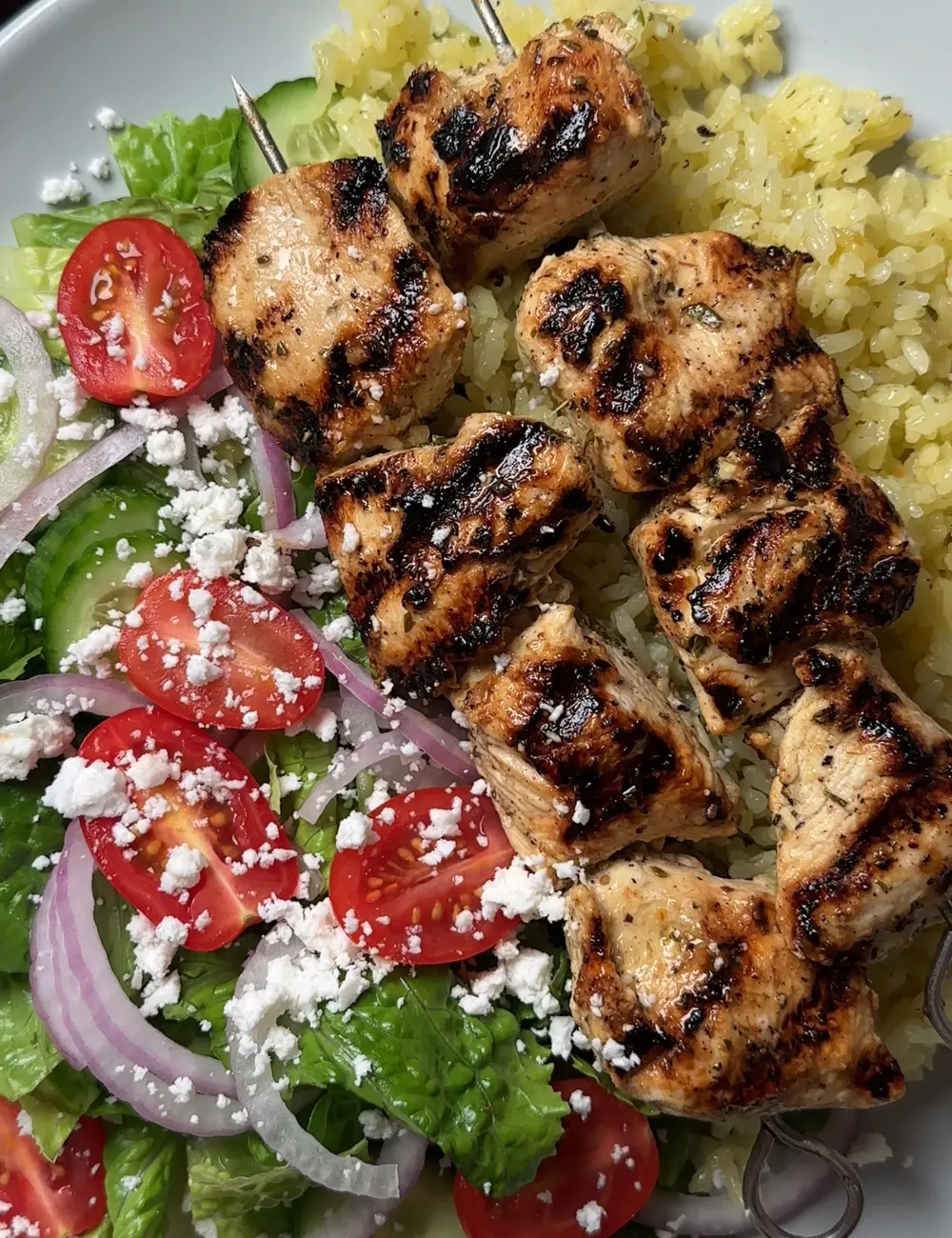 Grilled chicken skewers on yellow rice beside a Greek salad with tomato, cucumber, red onion and feta